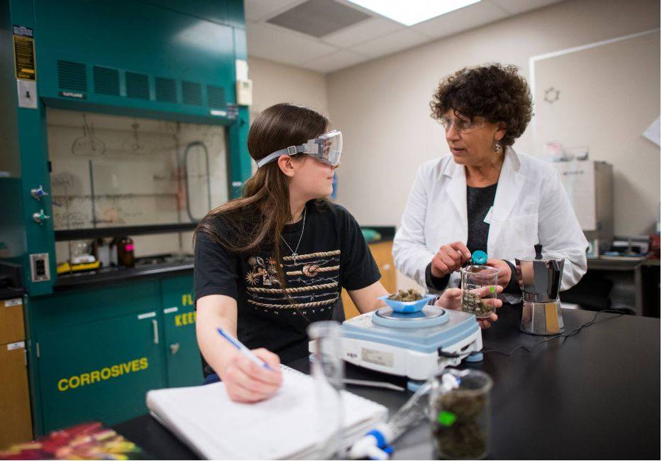 Student in goggles looks to instructor in a lab coat for guidance while measuring substance